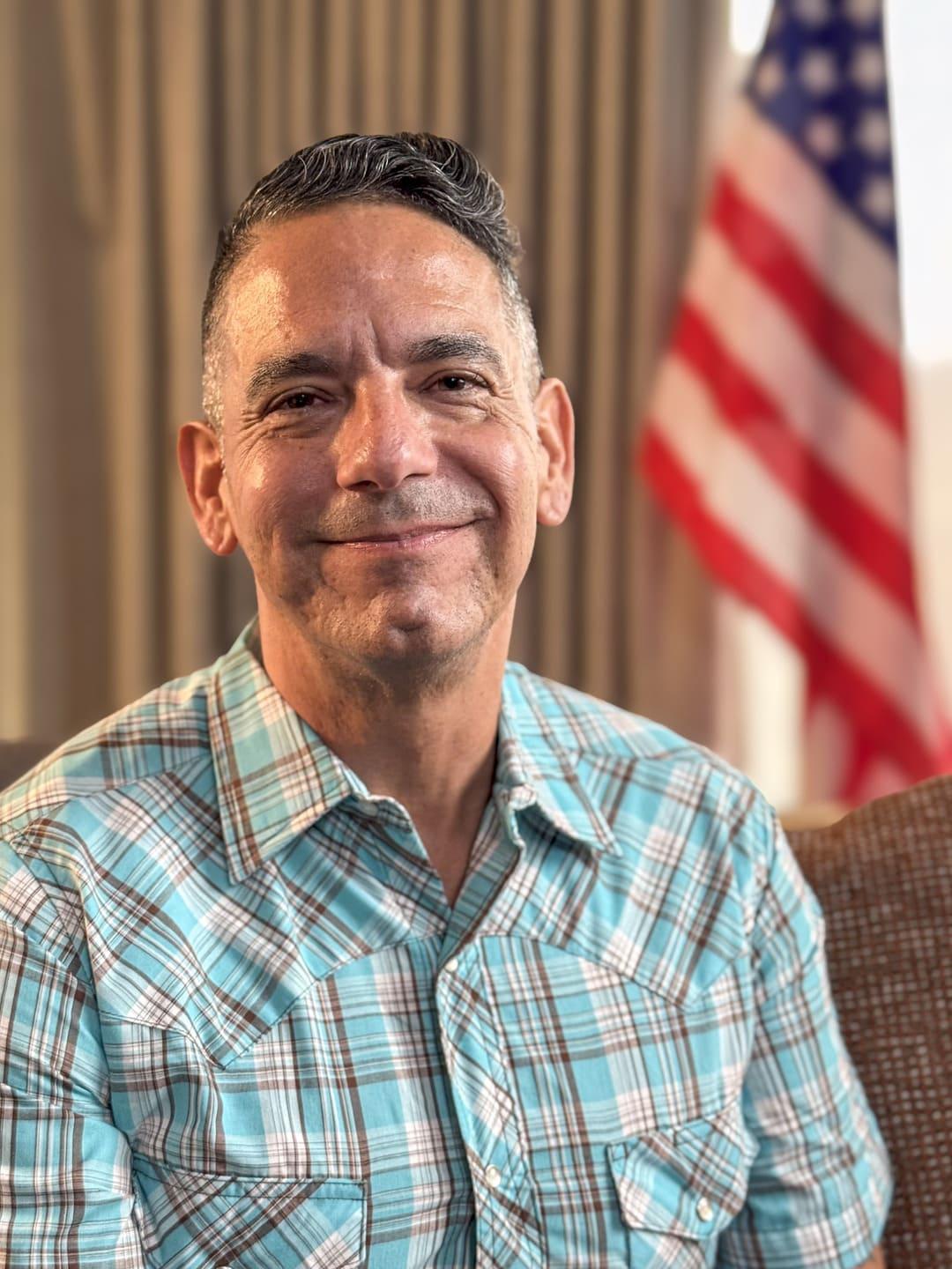 A man in a plaid shirt sits indoors with an American flag in the background.