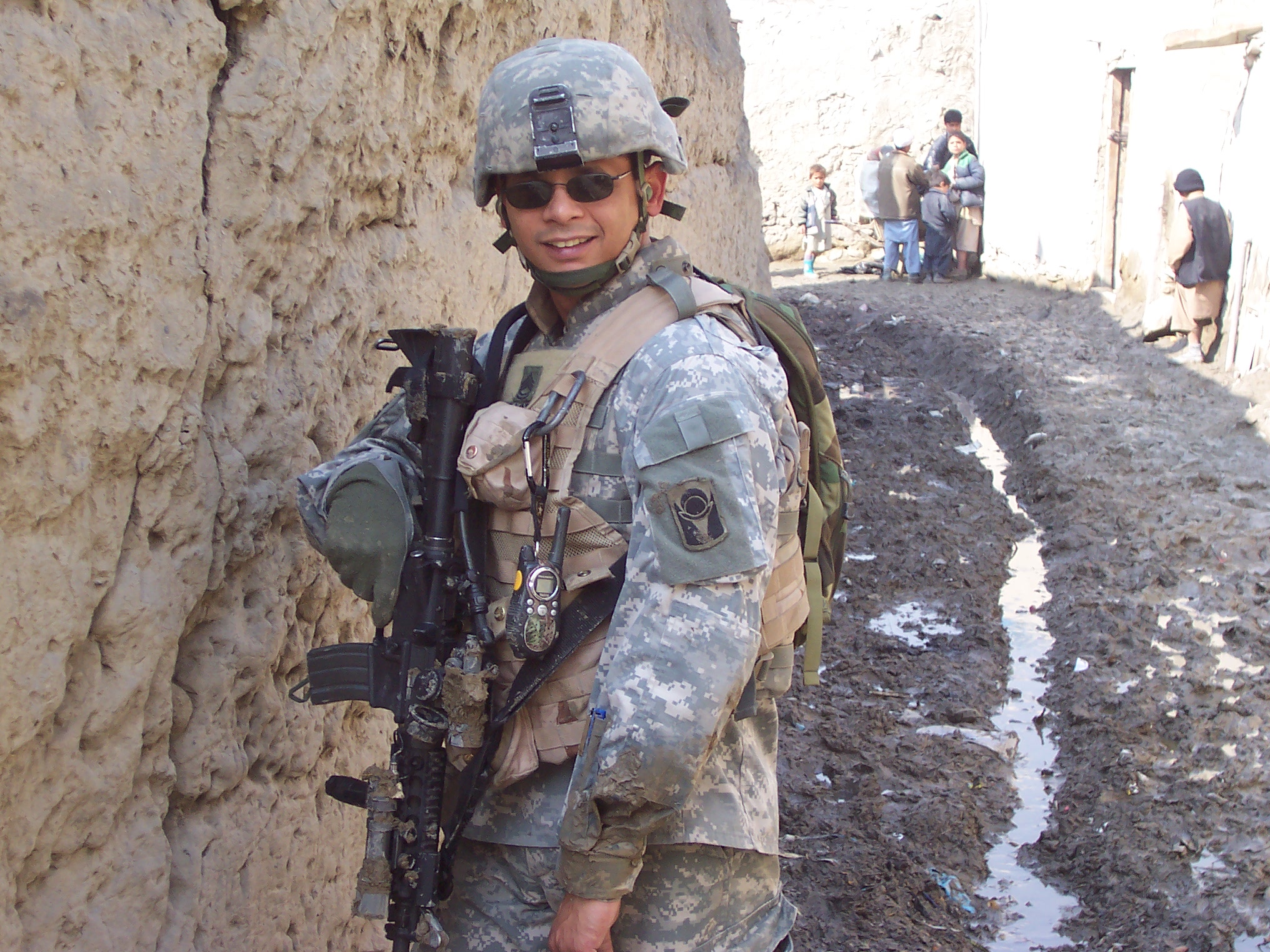 A soldier stands alert in a narrow dirt alley, surrounded by weathered walls and debris.