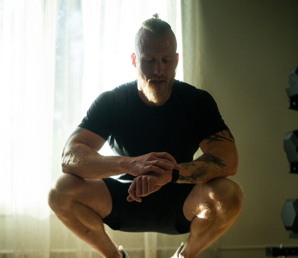 A man in workout clothing squats indoors while checking a smartwatch.